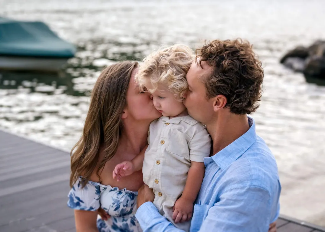 A family on a lake dock kissing their toddler
