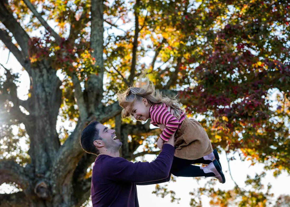 A father lifting his daughter into the air beneath an autumn tree