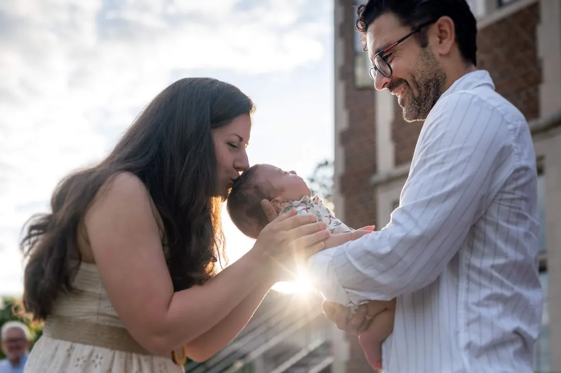 Parents leaning together to kiss their newborn in backlit window light