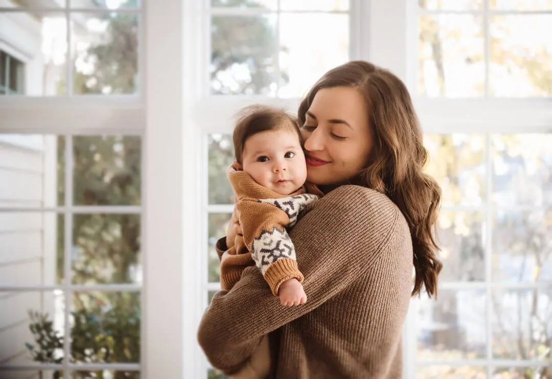 A mother holding her baby beside a bright window