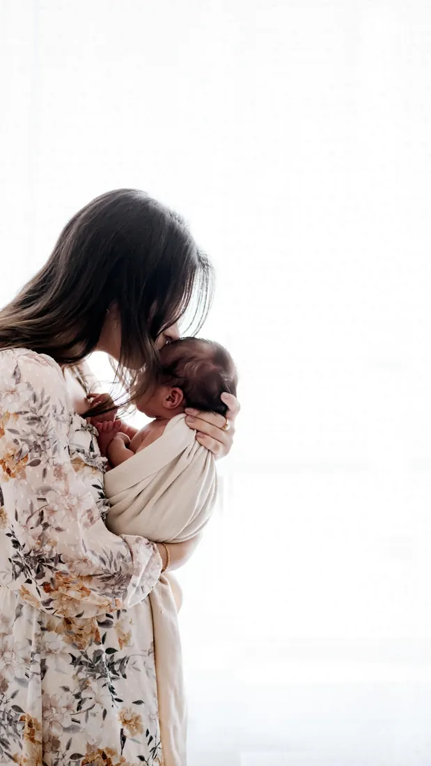 A mother kissing her newborn wrapped against her chest in soft window light